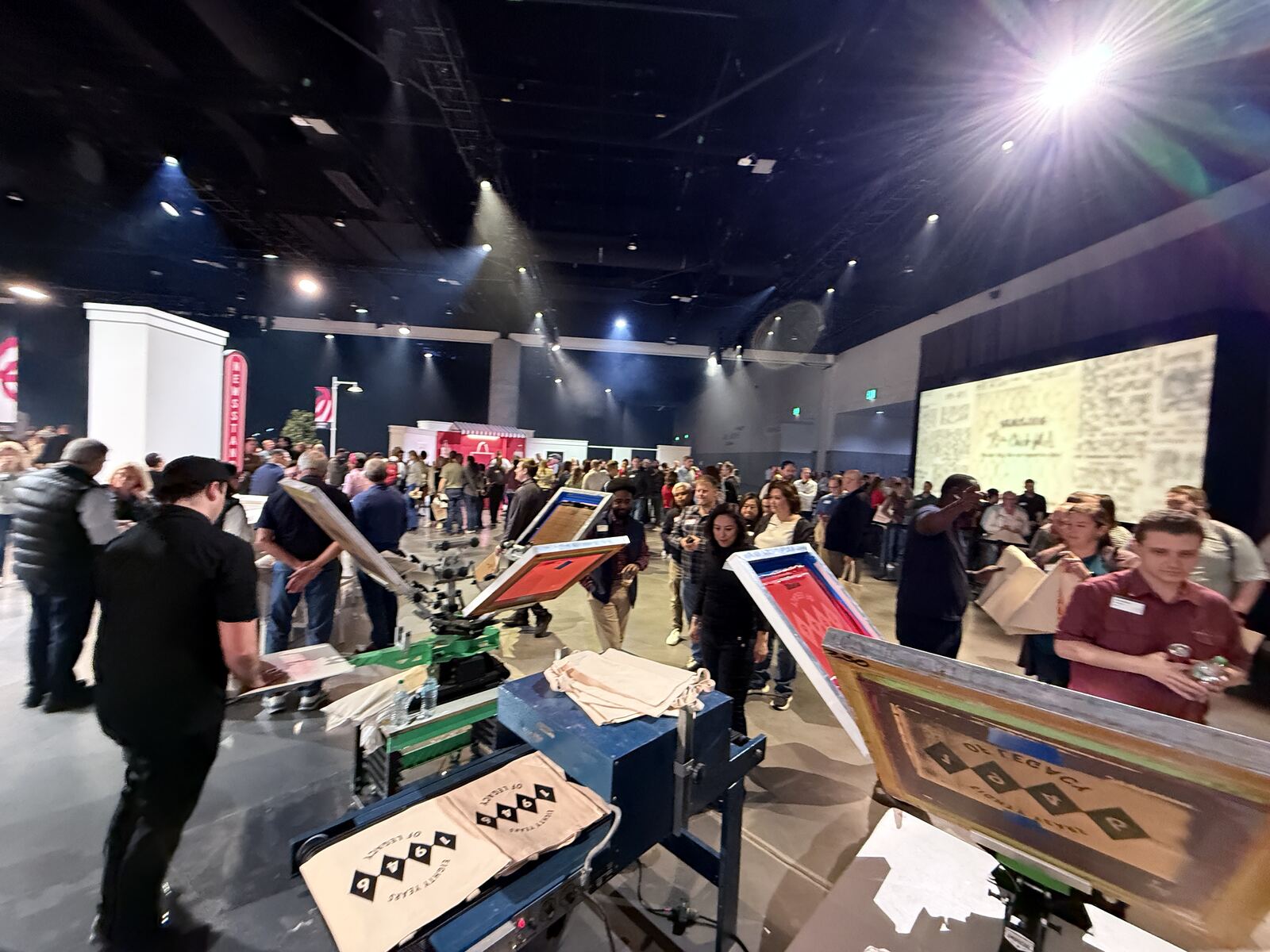 Vendor booth with a staff member and attendees browsing stacked apparel merchandise and branded products